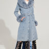 Modeling the KAYA FUR TRIM TRENCH IN DENIM, a person stands wearing this long trench coat featuring a faux fur collar, cuffs, belted waist, and sherpa lining. A skirt crafted from neckties flows beneath against a plain light-colored backdrop.