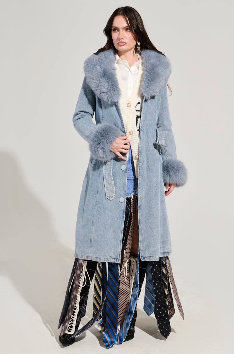 A woman stands confidently in the Kaya Fur Trim Trench in Denim. Beneath, she wears a white shirt and a tie-detail skirt featuring various patterns and colors. Her long dark hair cascades as she poses against a plain backdrop.