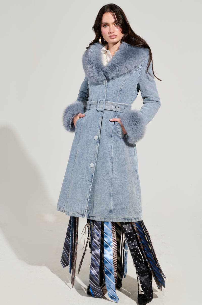 A woman in the KAYA FUR TRIM TRENCH IN DENIM, complete with a faux fur collar, stands with hands in pockets. She pairs it with a unique skirt crafted from colorful ties against a simple, neutral background.