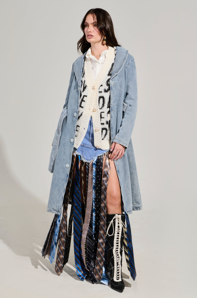 A woman models the KAYA FUR TRIM TRENCH IN DENIM, a light blue denim trench coat with a faux fur collar, an off-white sweater with black text, a necktie skirt, and black knee-high boots. She has long dark hair and poses against a plain background.