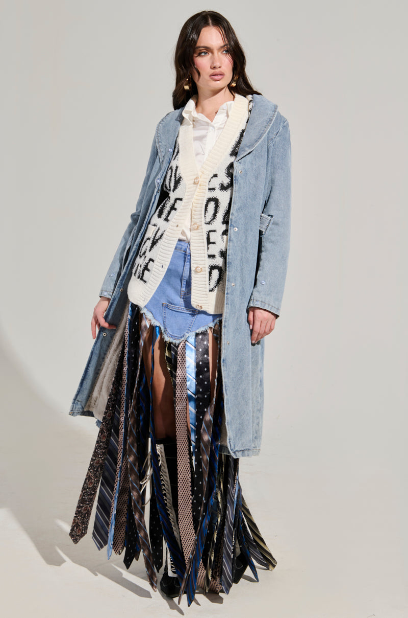A person wearing the KAYA Fur Trim Trench in Denim over a white shirt and patterned sweater. Their skirt is crafted from colorful, patterned neckties. The background is light gray.