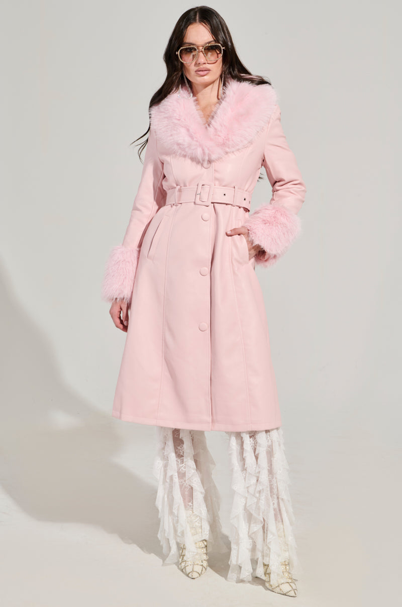 Dressed confidently in the KAYA Fur Lined Trench in Light Pink, a person showcases the coat's plush collar and cuffs. Its matching belt and button details perfectly enhance their sunglasses. Gracefully layered white lace pants and pointed shoes complete this chic ensemble.