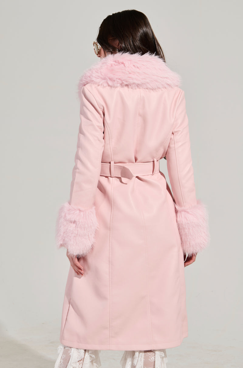 A person is wearing the KAYA Fur Lined Trench in Light Pink, featuring a fluffy collar and cuffs. They are facing away from the camera, highlighting the back of this faux leather coat with its belt. Their long hair cascades down as they stand indoors against a simple backdrop.