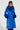 A person wearing the NEW KAYA FUR LINED TRENCH IN BLUE, featuring a long, vibrant blue faux leather design with fluffy faux fur lining on the collar and cuffs, stands against a plain background. Their hair is braided, and they sport an adjustable belt along with a confident expression.