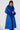 The individual with braided hair exudes confidence, showcasing the NEW KAYA FUR LINED TRENCH IN BLUE. This striking coat, with its fluffy cuffs and collar, is stylishly fastened using its adjustable belt as they pose against a plain white background.