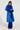 A person with braided hair stands confidently in the NEW KAYA FUR LINED TRENCH IN BLUE, flaunting fur details on the collar and cuffs over a long, textured dress. An adjustable belt cinches the waist, adding a flattering touch against the plain background.