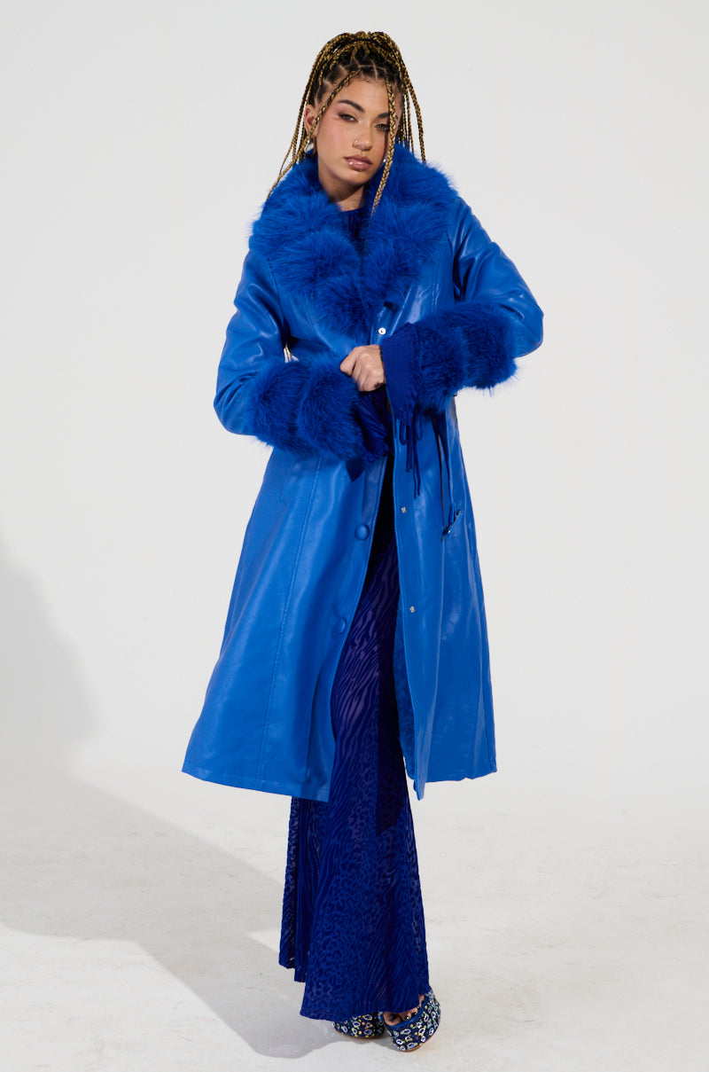 A person wearing the NEW KAYA FUR LINED TRENCH IN BLUE and a textured blue dress stands against a plain background. They have braided hair and are sporting leopard-print shoes.