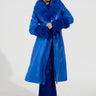 A person wearing the NEW KAYA FUR LINED TRENCH IN BLUE and a textured blue dress stands against a plain background. They have braided hair and are sporting leopard-print shoes.