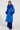 A woman exudes confidence in the NEW KAYA FUR LINED TRENCH IN BLUE, with its vibrant faux leather finish and plush collar and cuffs. Her hands rest comfortably in its pockets, perfectly complementing her long, textured blue skirt. Her hair is styled in cornrows against a plain white background.