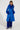 Wearing the chic NEW KAYA FUR LINED TRENCH IN BLUE, a person stands confidently with the jacket's fluffy cuffs and collar accentuating their look. The trench is layered over a textured blue dress, complementing their braided hairstyle as they glance slightly to the side against a plain white background.