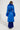 Rear view of a person wearing the NEW KAYA FUR LINED TRENCH IN BLUE with a belt. They have braided hair and are dressed in a long skirt with matching boots, standing against a plain background.