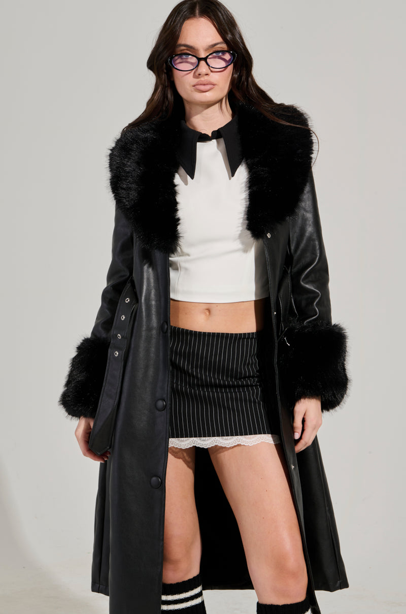 A person is seen wearing glasses and the KAYA FUR TRIM TRENCH, a black coat with faux fur detailing, layered over a white top. This chic ensemble is paired with a pinstriped skirt that features an adjustable waist belt and lace hem, all beautifully complemented by knee-high socks for a stylish modern look with contrasting textures.