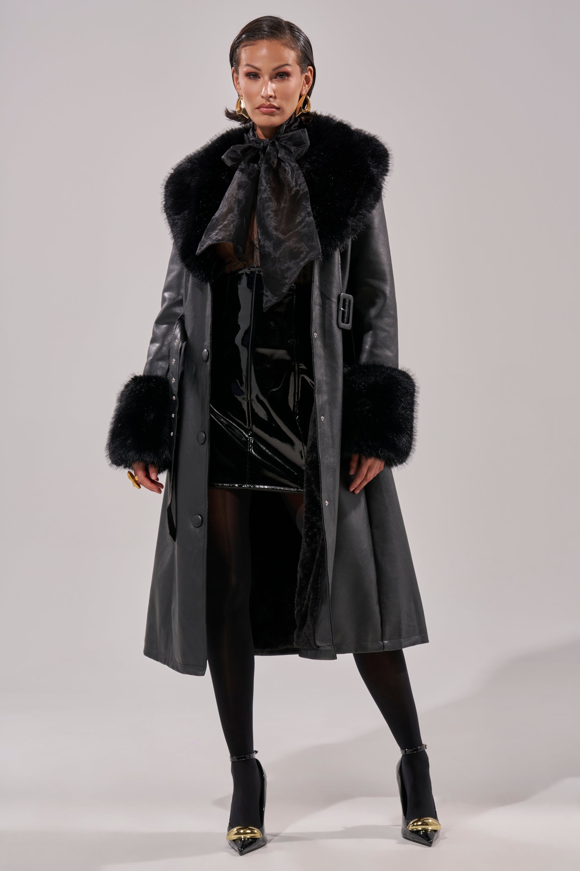A woman stands confidently in the KAYA FUR TRIM TRENCH with fur accents on the collar and cuffs, paired with a shiny black mini dress, opaque tights, and black heels featuring gold toes.