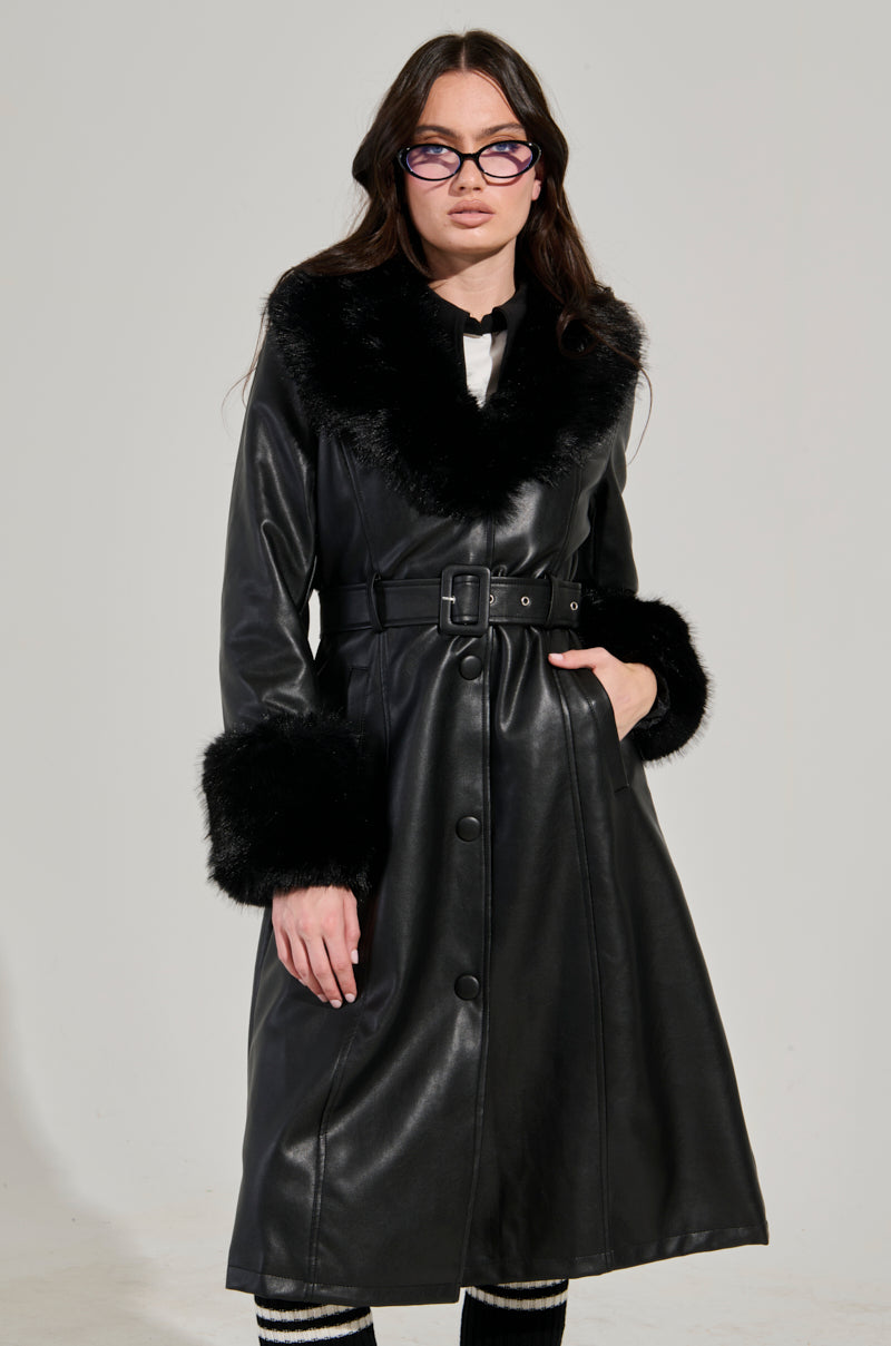 A person wearing the KAYA FUR TRIM TRENCH, a black leather coat with faux fur accents on the collar and cuffs, stands against a plain, light-colored background. The coat is buttoned up and includes an adjustable waist belt. The person is also wearing glasses.