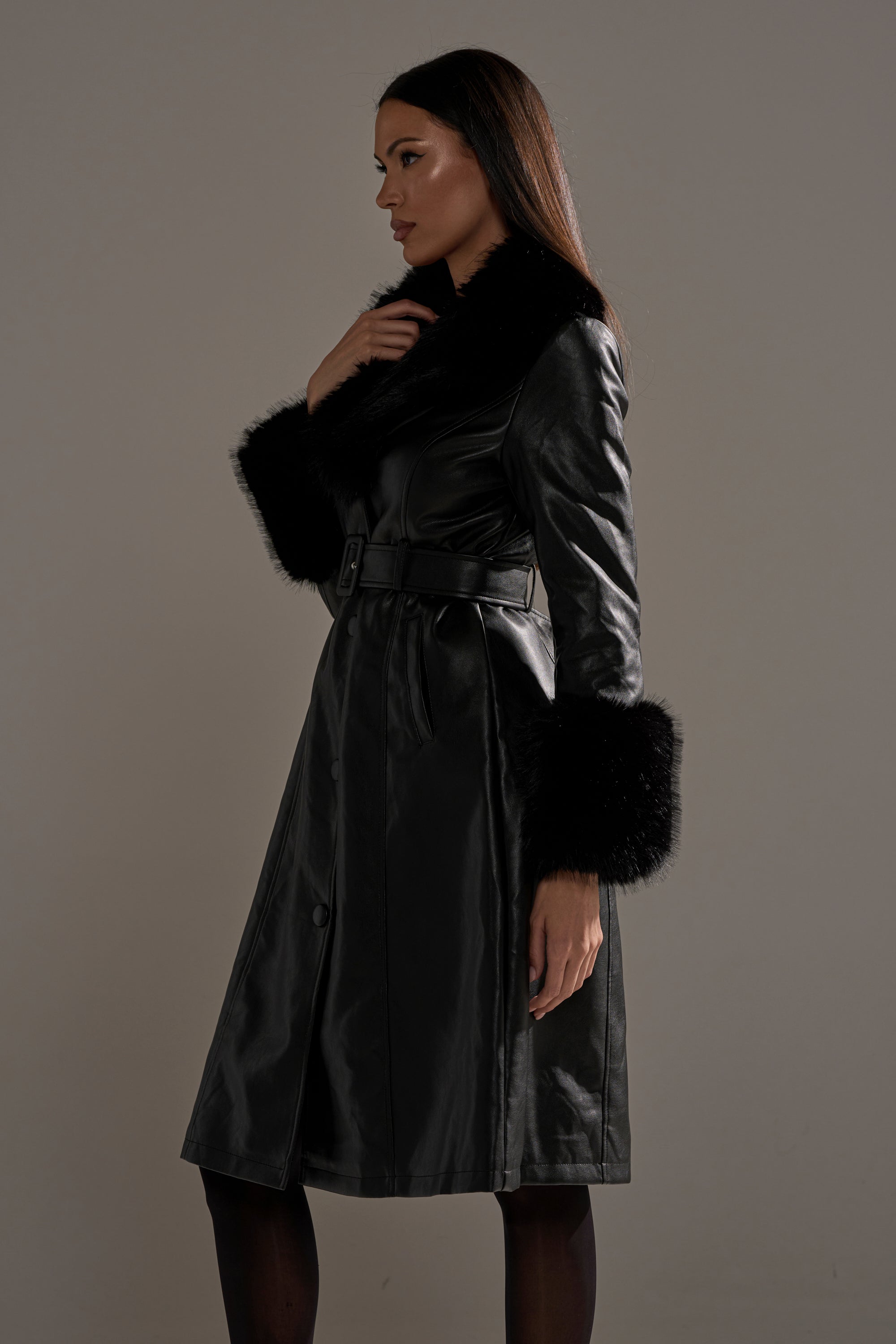 Woman in the KAYA FUR TRIM TRENCH—a black pleather coat with faux fur collar and cuffs—stands in profile against a neutral background, one hand touching her collar as she looks ahead.