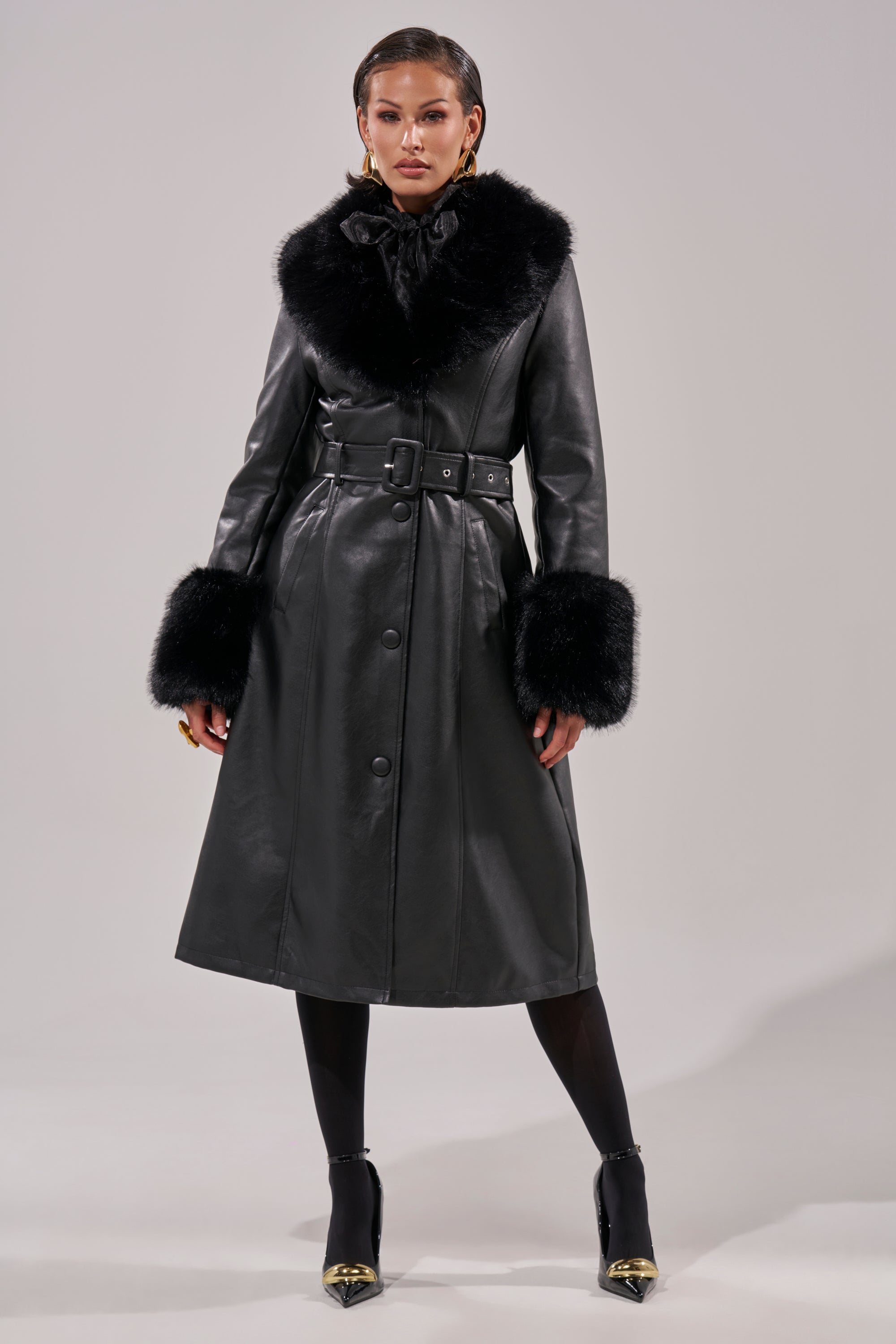 A woman models the KAYA FUR TRIM TRENCH, a black pleather coat with a wide belt and faux fur on the collar and cuffs, styled with black tights and pointed gold-tip heels against a plain light background.