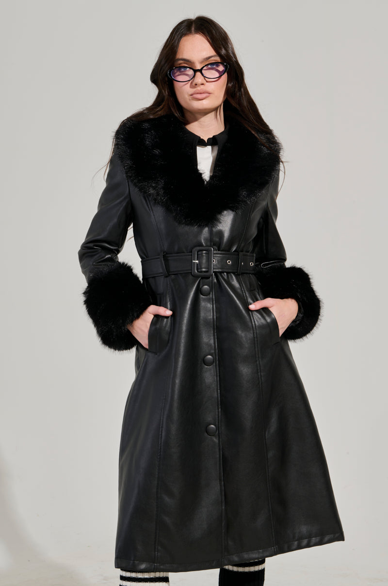A person dressed in the KAYA FUR TRIM TRENCH, a long black pleather coat with a faux fur collar and cuffs, is standing with hands in pockets. They have long dark hair and are wearing glasses. The coat includes an adjustable waist belt for a perfect fit.