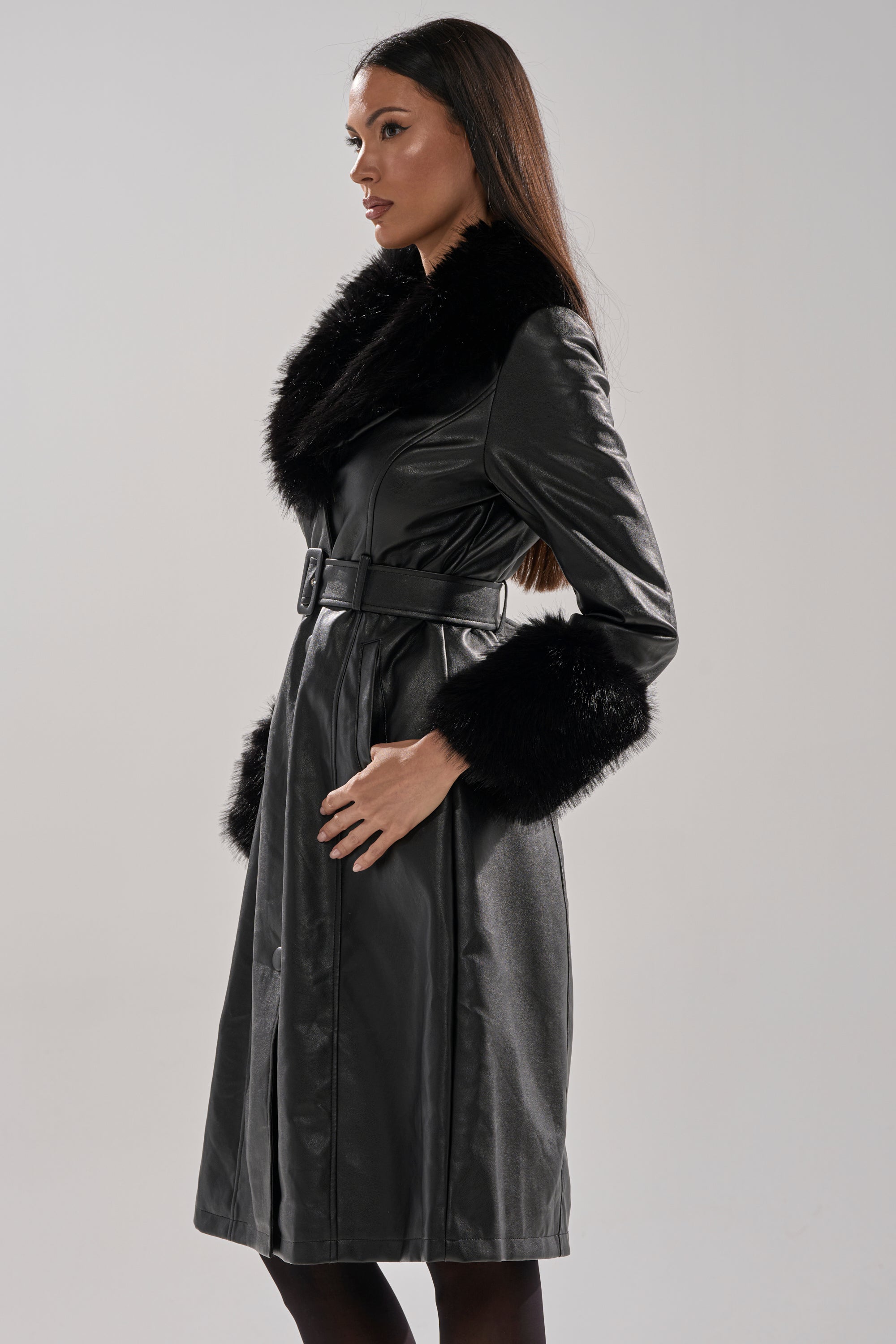 A woman stands in profile wearing the KAYA FUR TRIM TRENCH, a black zip-up coat featuring a wide faux fur trim collar and cuffs, with hands in pockets, long straight hair, and a neutral background.