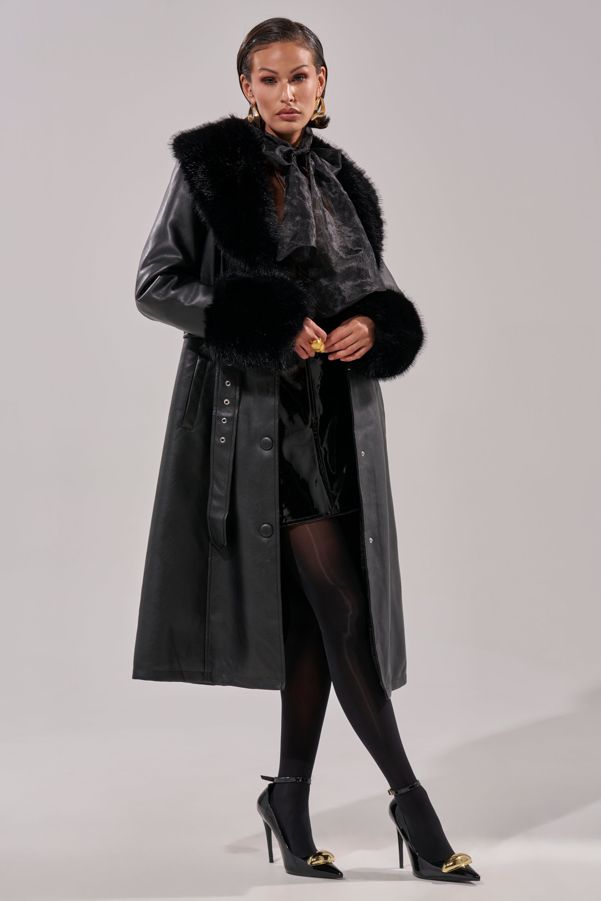 A woman poses against a plain gray background wearing the KAYA FUR TRIM TRENCH, styled with sheer black tights, black high heels featuring gold details, and gold hoop earrings.