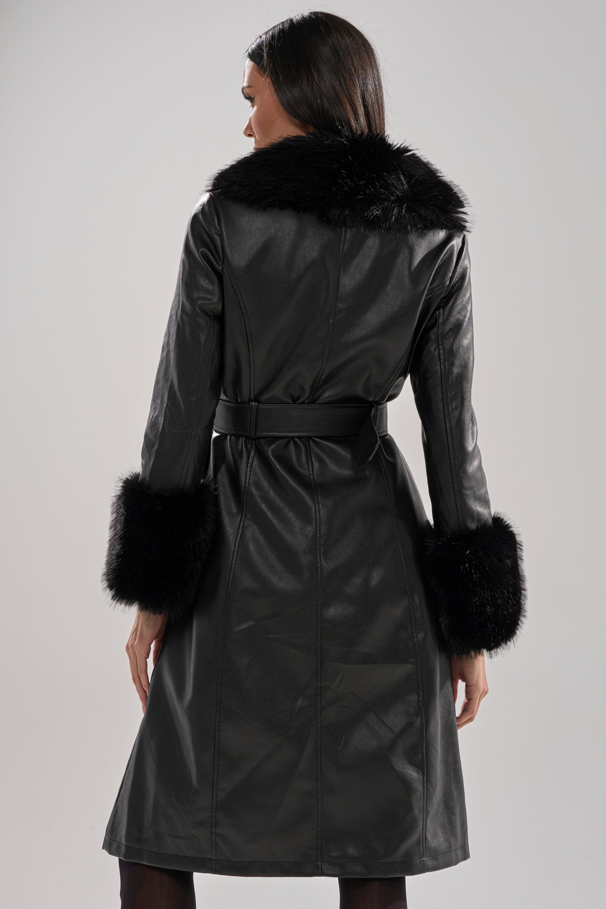 A woman with long dark hair is seen from behind wearing the KAYA FUR TRIM TRENCH, a long black zip-up coat featuring faux fur trim on the collar and cuffs, set against a plain light background.