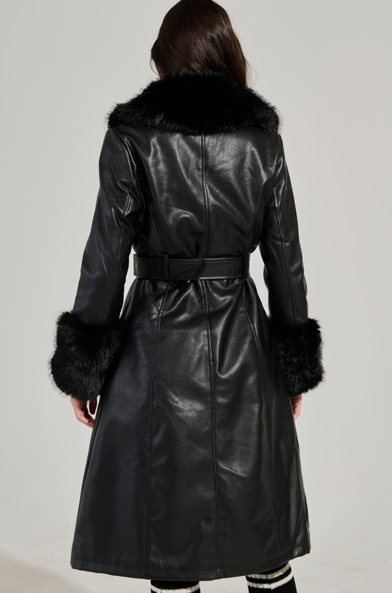 A person wearing the KAYA FUR TRIM TRENCH stands with their back to the camera. The coat, featuring a faux fur trim collar and cuffs, is cinched at the waist with an adjustable belt, perfectly complementing their black and white striped pants against a plain gray background.