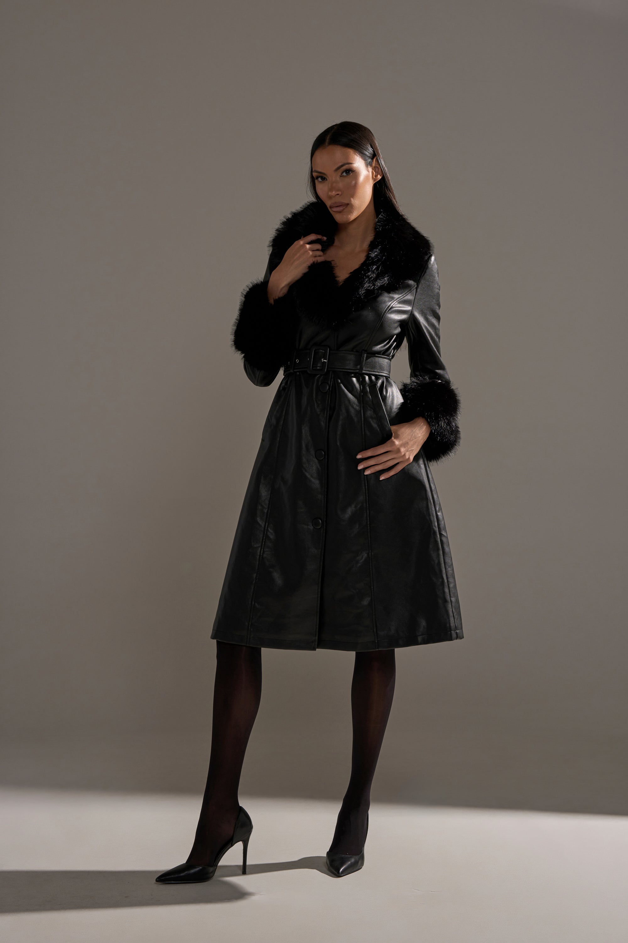 A woman stands confidently in the KAYA FUR TRIM TRENCH, a black zip-up coat with faux fur collar and cuff accents, paired with black tights and heels against a softly lit background.
