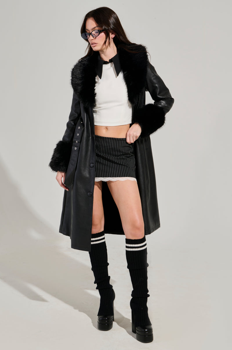 A person stands confidently in the KAYA FUR TRIM TRENCH, a black pleather zip-up coat with an adjustable waist belt, featuring a faux fur collar and cuffs. The ensemble is complemented by a white top, a black skirt, and knee-high boots with stripes. With long hair and glasses, they gaze slightly to the side.