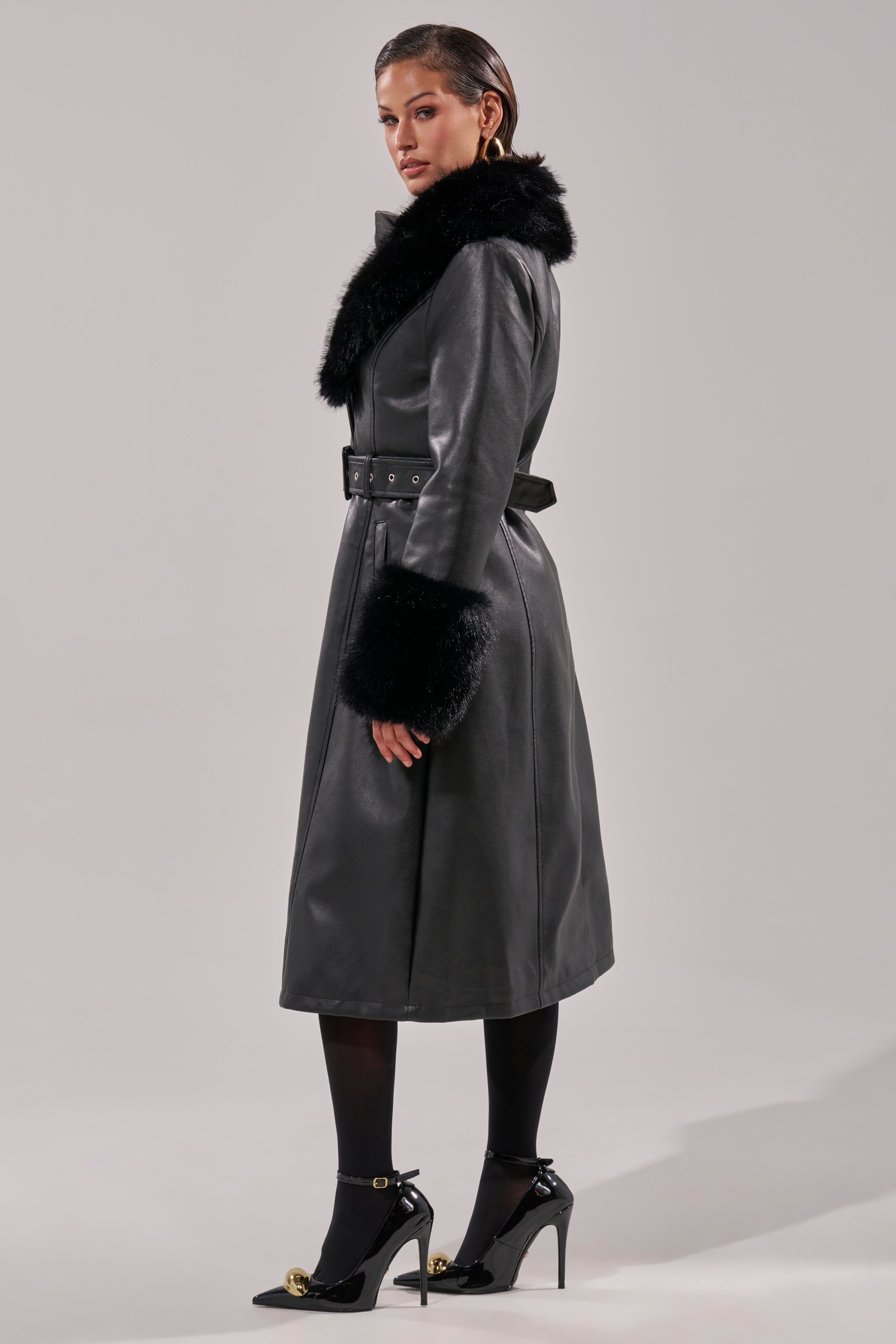 A woman stands in profile wearing the KAYA FUR TRIM TRENCH, a long belted pleather coat with black faux fur at the collar and cuffs, styled with black tights and high heels against a simple, neutral background.