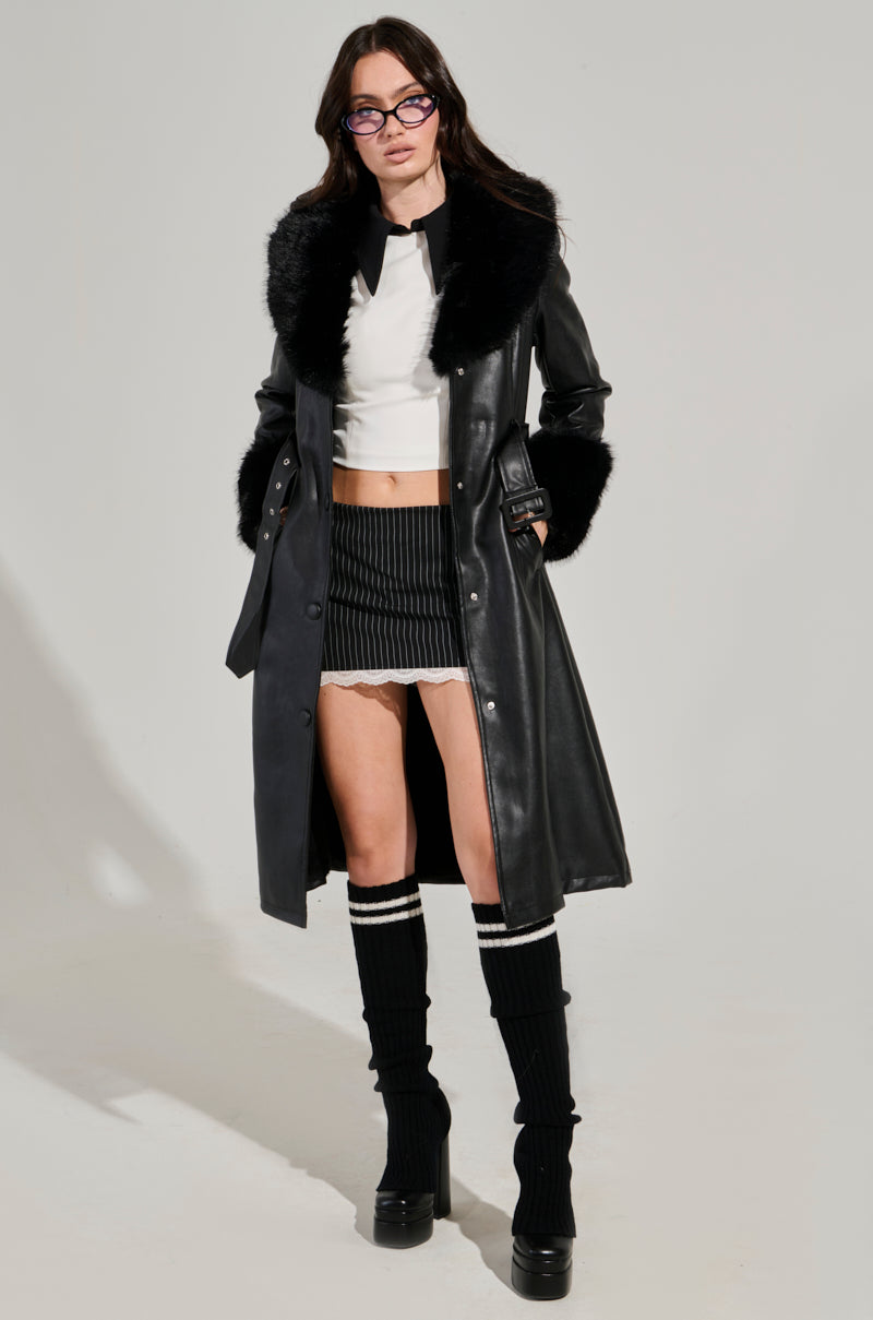 A person is wearing the stylish KAYA FUR TRIM TRENCH over a white cropped top and a black pinstripe mini skirt. Knee-high black socks with white stripes peek out above platform shoes, while glasses add a touch of sophistication.
