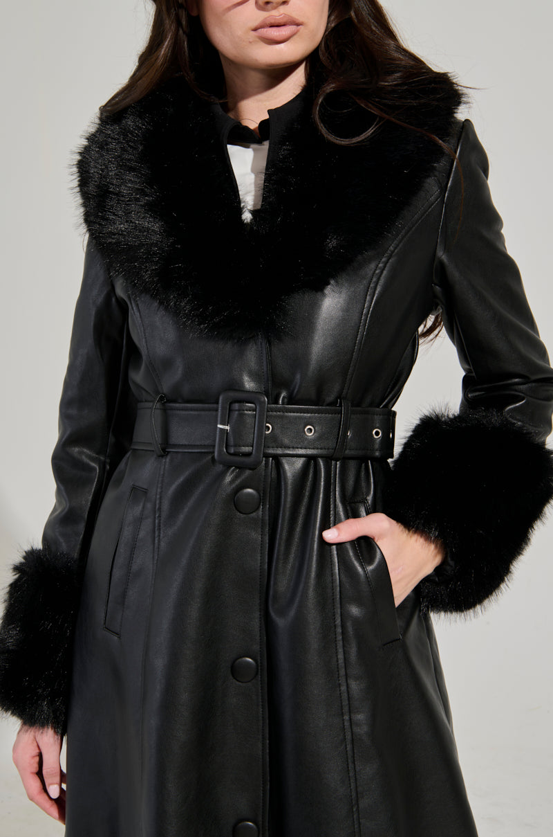 A person is wearing the KAYA FUR TRIM TRENCH, a black leather coat with a wide faux fur trim collar and cuffs. This stylish trench coat is belted at the waist for an adjustable fit and features button details. The background is plain and light-colored.