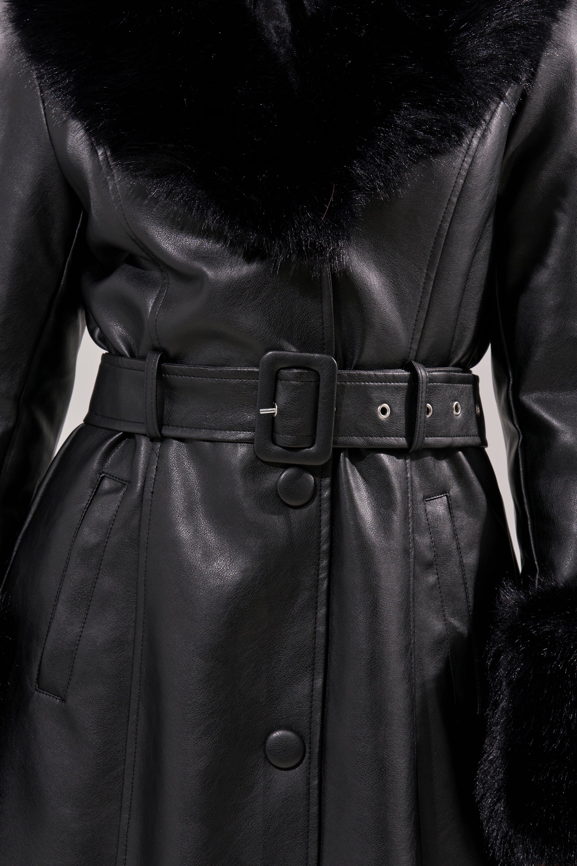 The KAYA FUR TRIM TRENCH is a black pleather coat with wide faux fur trim at the collar and cuffs, snap buttons, a matching belt with square buckle, and pockets where the wearer’s hands are partially visible.