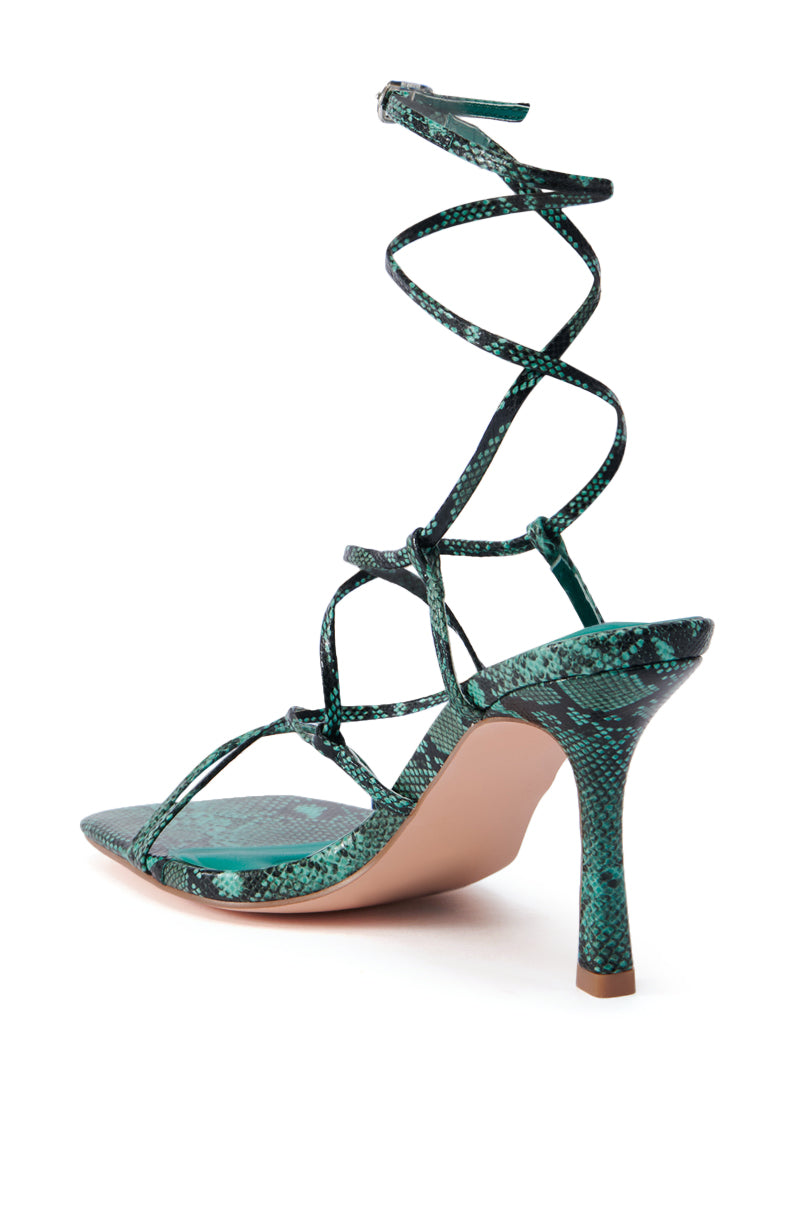 Introducing the AZALEA WANG KAYLIN GREEN SNAKE SANDAL, a stunning high-heeled shoe featuring green snake-print straps and a slender stiletto heel. This design showcases multiple thin straps that elegantly cross over the foot and wrap around the ankle. The chic look is completed with a light beige sole, offering a graceful view from the back at an angle.