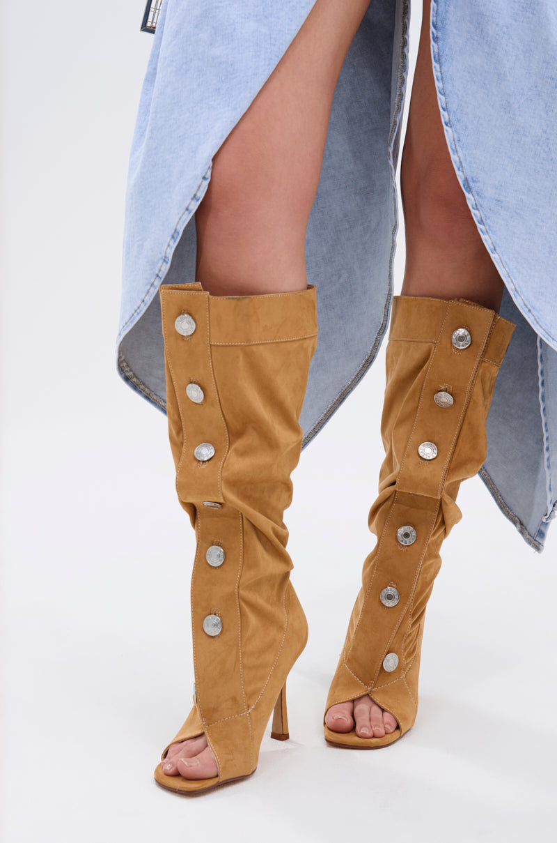 A person wears the AZALEA WANG KAZUKI TAN SLOUCHY SUEDE OPEN TOE BOOT—tan, knee-high, with large silver buttons and a stiletto heel—paired with a light blue, high-slit denim skirt against a white, minimal background.