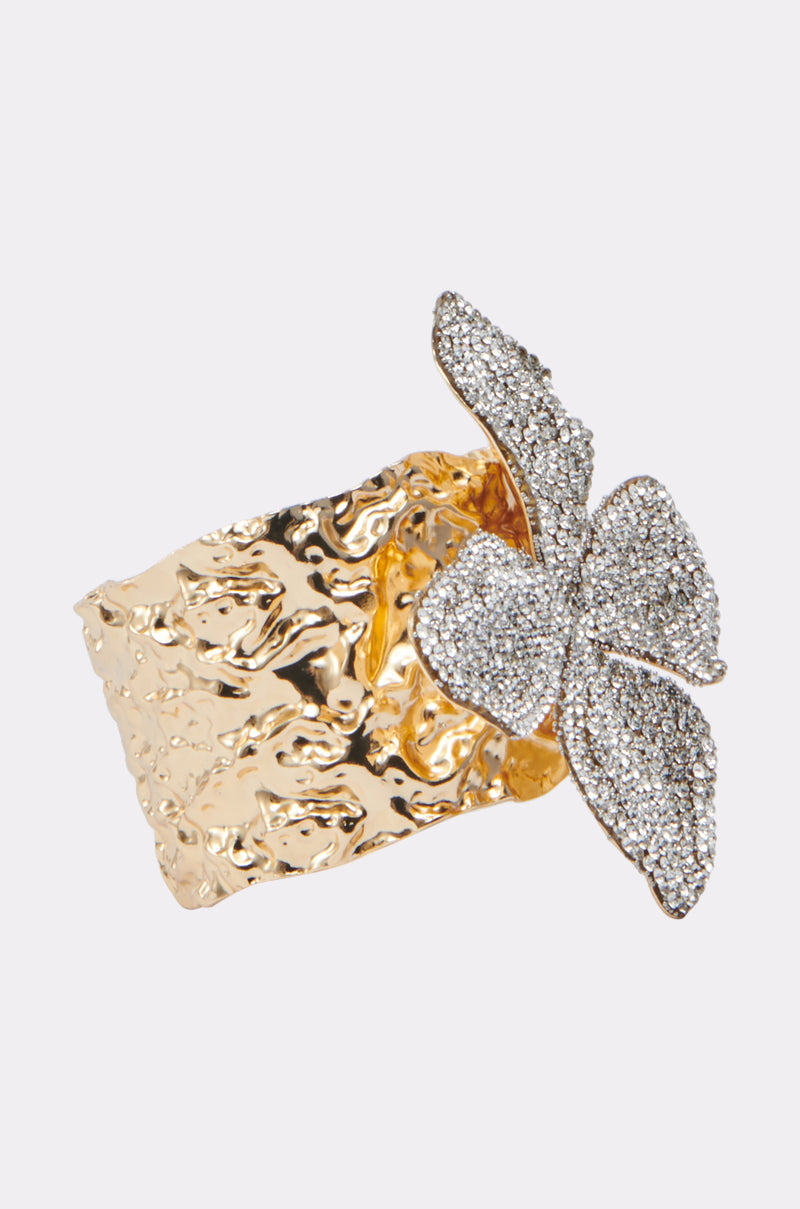 The EMI FLOWER BRACELET by AKIRA Label is a wide, hammered gold-tone cuff featuring a large crystal-encrusted butterfly design, shown on a plain white background. 2