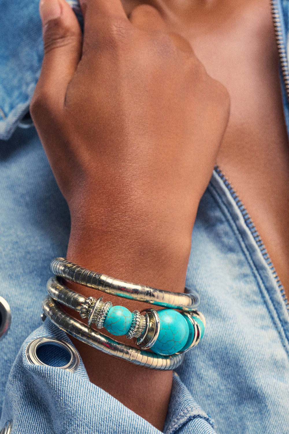 Wearing a light denim jacket with the sleeves rolled up, a person shows off boho-chic jewelry, including silver bangles and the SANTORINI SUNSET BRACELET, which features striking turquoise accents on their wrist.