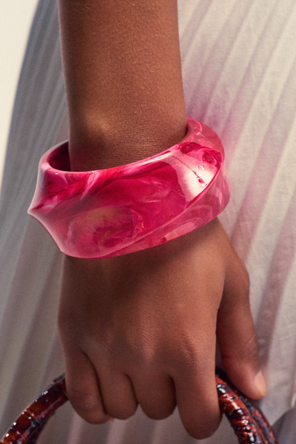 A close-up of a person's hand adorned with the Resin Swirl Bangle in Pink—a true piece of statement jewelry—holding a reddish-brown textured handle, with a pleated white skirt in the background.