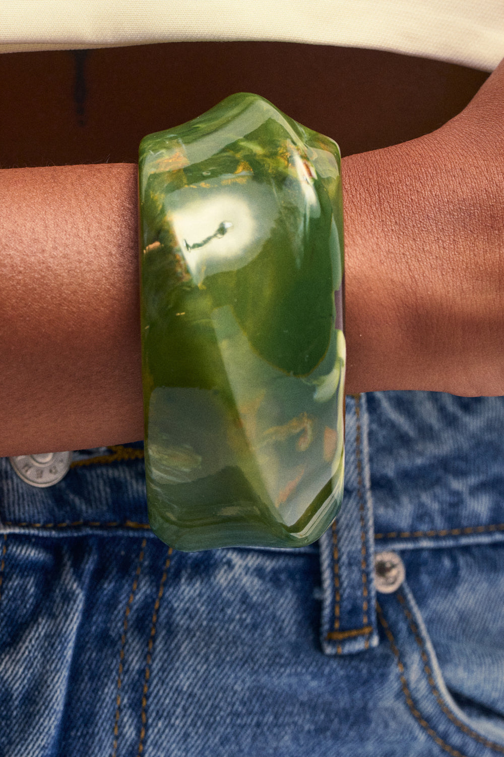 A person wearing the RESIN SWIRL BANGLE IN GREEN—a chunky, faceted green marbled resin bracelet—on their wrist, shown against tan skin and blue denim jeans. This sculptural accessory adds a touch of boho-chic jewelry flair to the look.