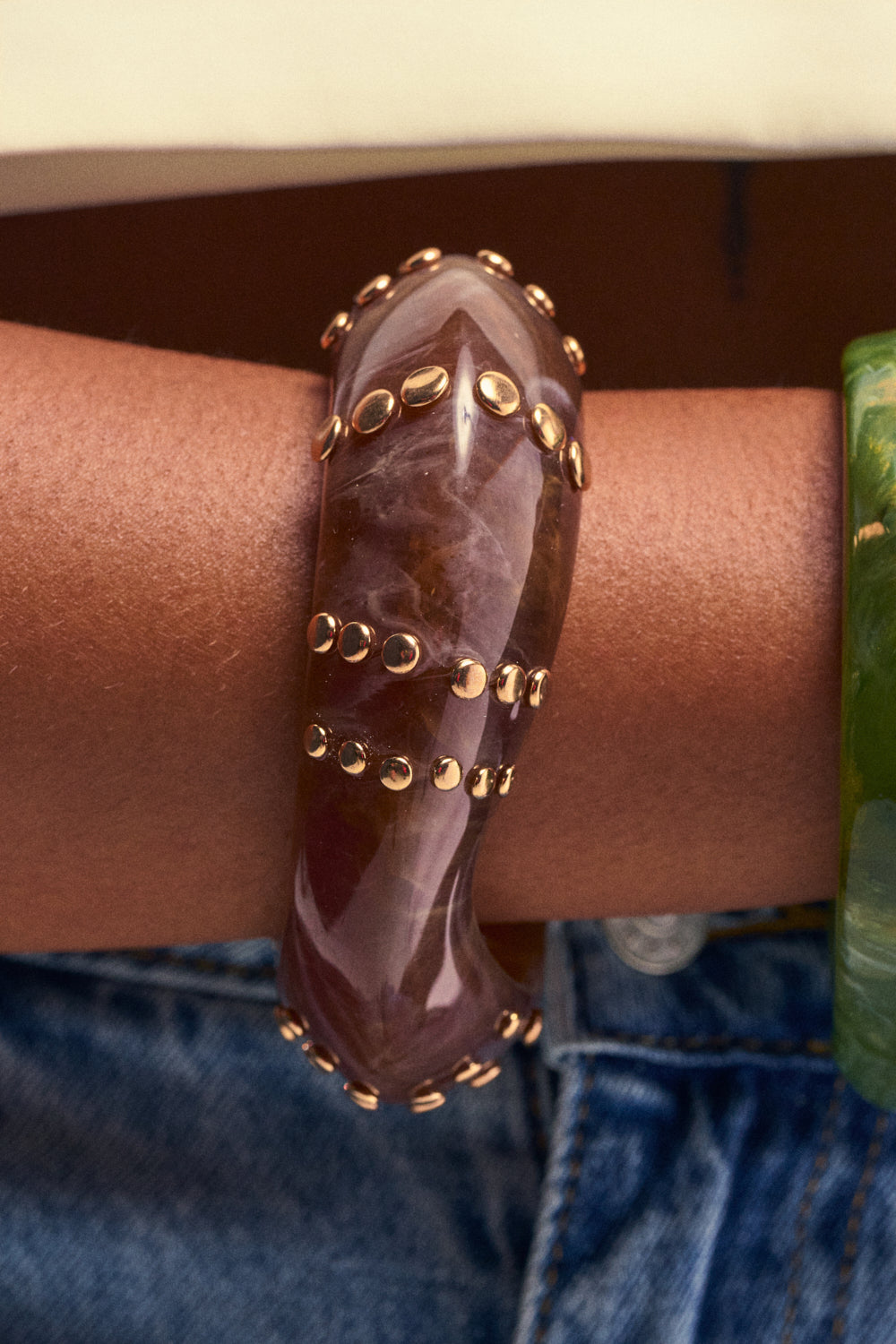 A close-up of a wrist accessorized with a chunky, curved, purple stone boho bracelet accented by metallic studs, paired with the MOCHA GLOW RESIN BRACELET in green. The background features denim jeans and a glimpse of a beige shirt.