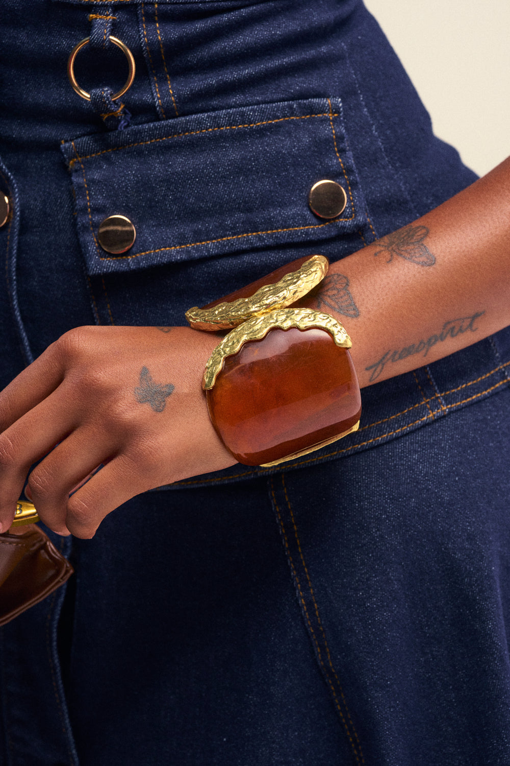 Wearing a dark denim outfit, a person shows off their butterfly and "transport" tattoos while accessorizing with the SWEET LIKE HONEY BUTTERFLY CUFF—a striking acrylic bracelet featuring gold hammered accents, perfect for boho summer jewelry lovers.