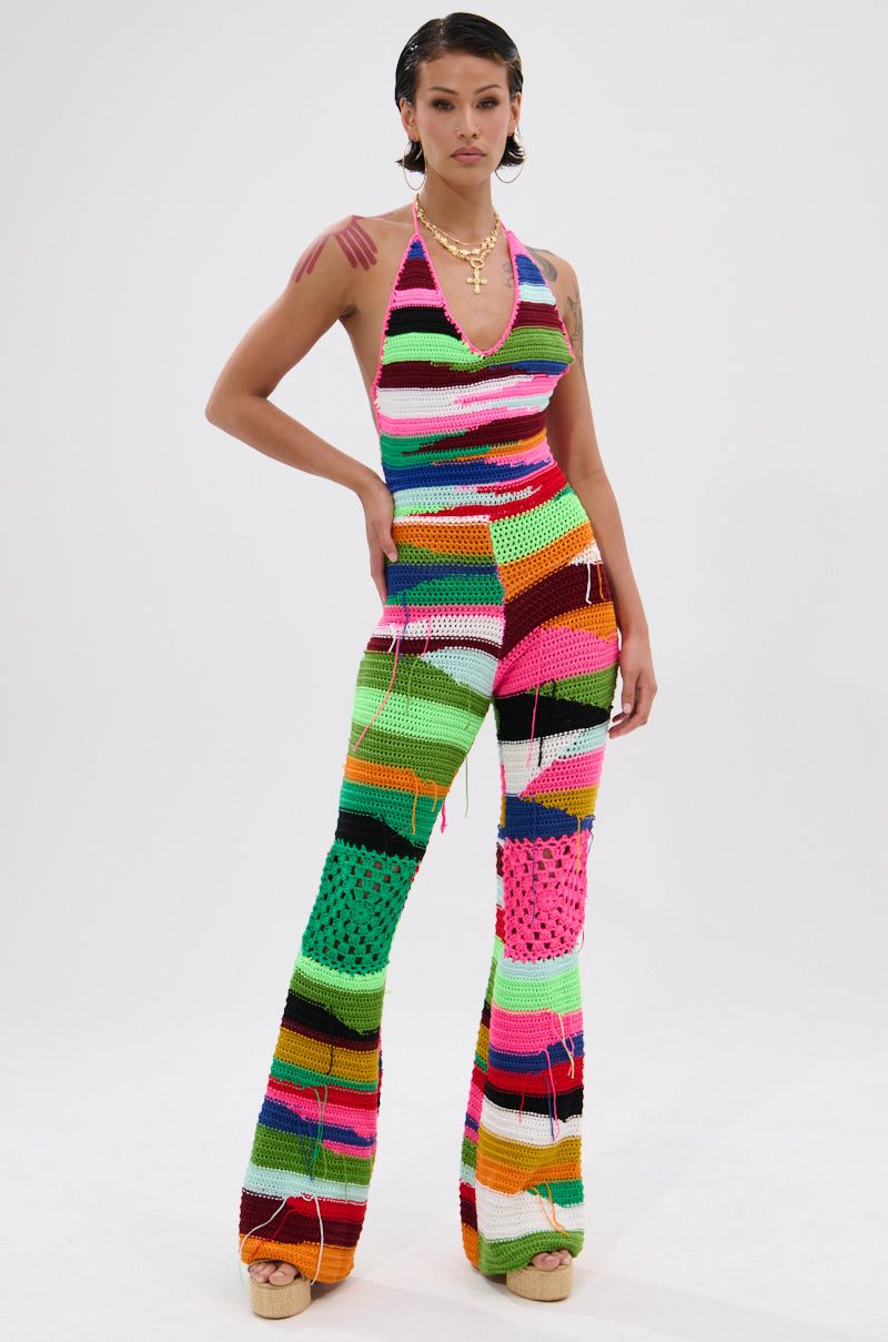 A person stands confidently in the FUNKY TOWN CROCHET JUMPSUIT, a flared-leg piece with bold patchwork patterns and vibrant colors. Its striped halter design pops against the plain white background.