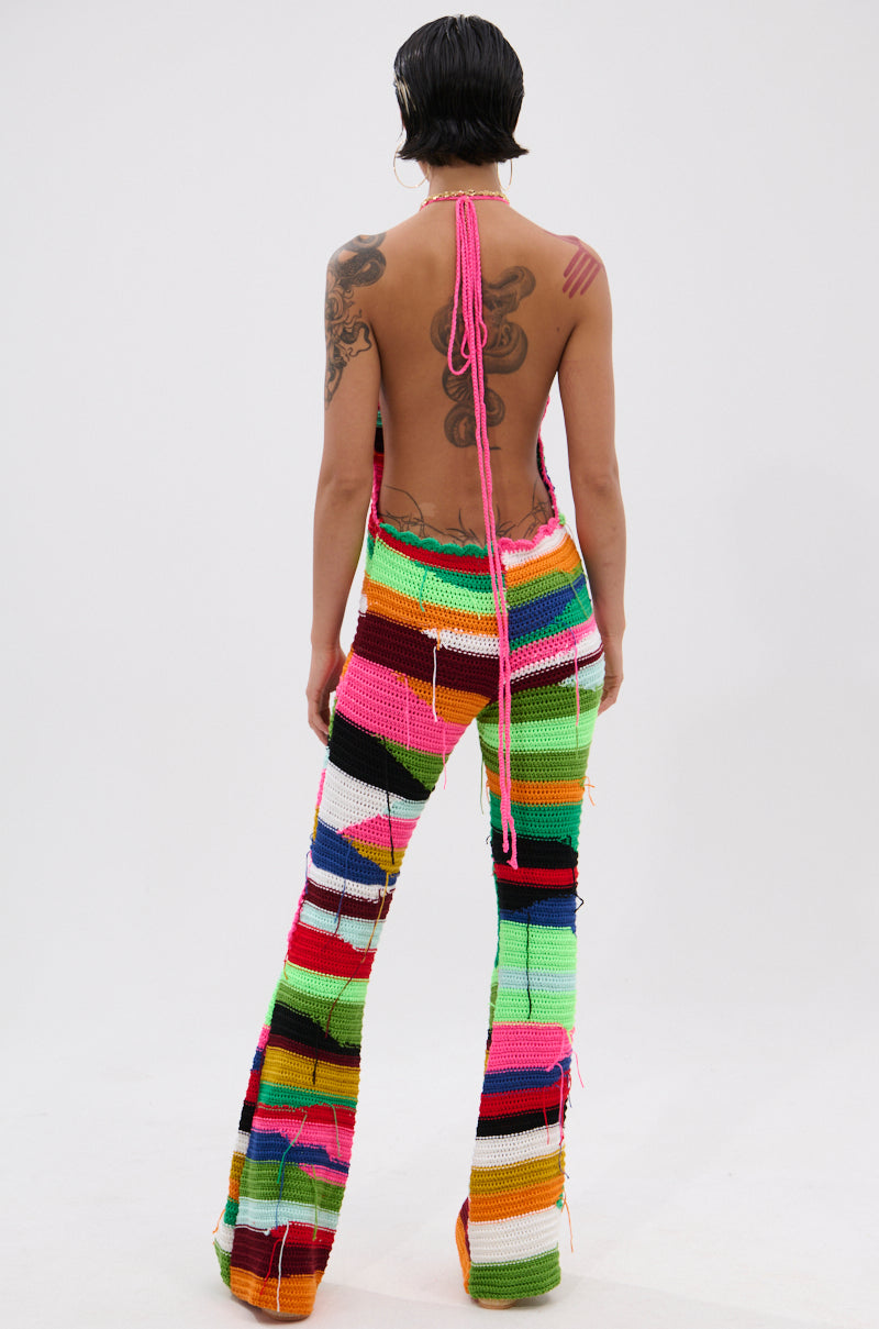 A person with short dark hair and visible tattoos stands facing away against a plain white background, wearing the FUNKY TOWN CROCHET JUMPSUIT featuring vibrant colors, flared legs, a backless halter top, and loose threads.