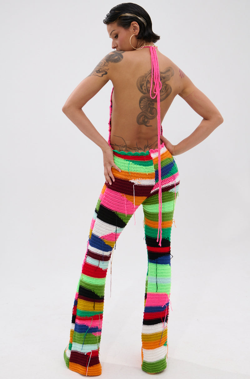 A person faces away in the FUNKY TOWN CROCHET JUMPSUIT, featuring bright pink, green, orange, yellow, and black stripes. The flared legs and halter top with an open back showcase bold tattoos beneath the vibrant crochet design.