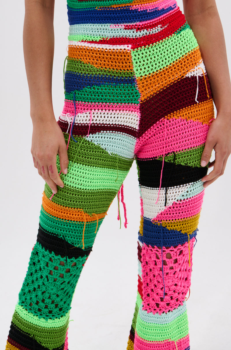 A person models the FUNKY TOWN CROCHET JUMPSUIT, featuring colorful crocheted patchwork, loose yarn details, and a flared leg silhouette. Only the lower torso and upper legs are shown against a plain white background.