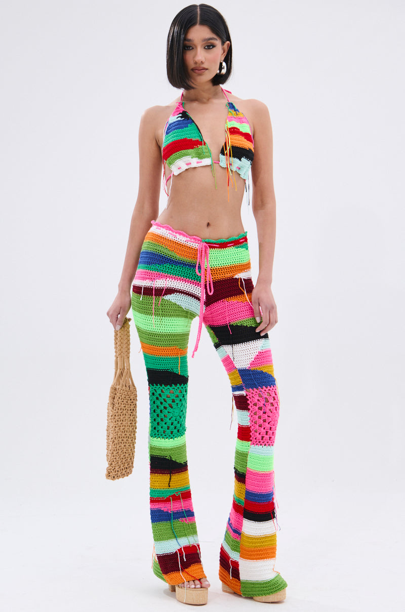 A woman poses against a white background in the MULTI COLORED HEART CROCHET BRA TOP, paired with matching flared pants, holding a woven tan handbag and wearing beige platform sandals for a festival-ready look.