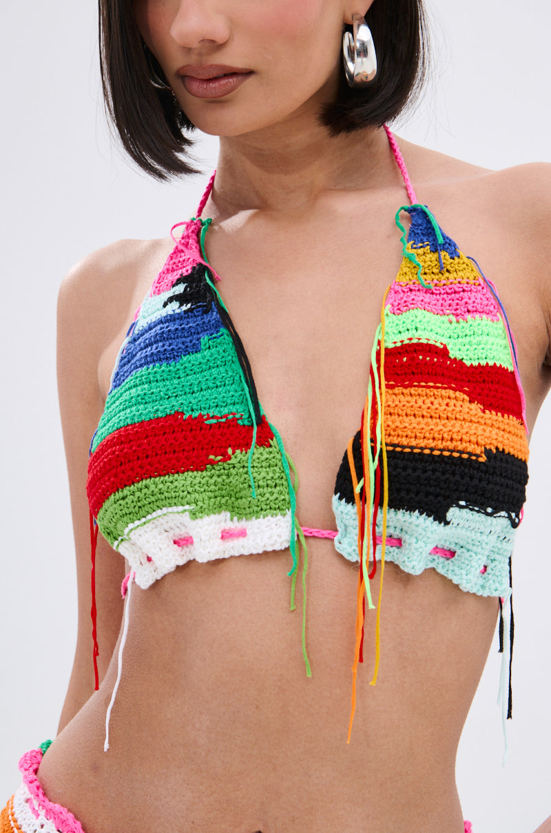 A woman wears large silver hoop earrings and the MULTI COLORED HEART CROCHET BRA TOP, featuring colorful stripes and loose multicolored yarn ends, shown from shoulders to waist against a plain background.