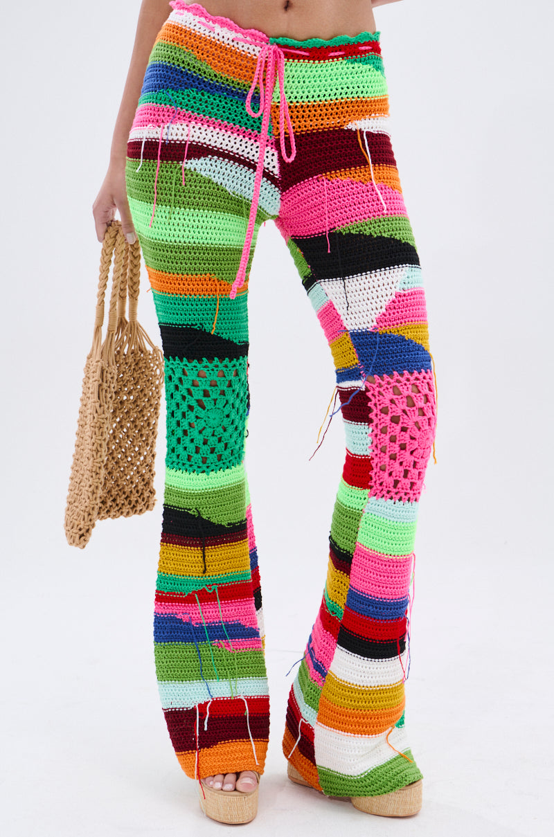 Against a plain white background, a person wears the COLORED HEART CROCHET PANT—high-waisted, fit-and-flare with vibrant multicolored stripes and heart patches—paired with a woven bag and platform sandals for a vintage-meets-avant-garde look.