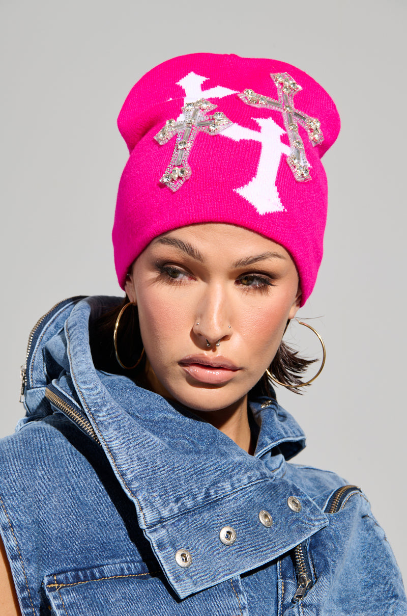 A person wears a vibrant CROSS LOVE BEANIE, featuring religiously-inspired crosses and silver rhinestones, paired with large hoop earrings and a blue denim vest. The plain gray background enhances this striking style even further. 0
