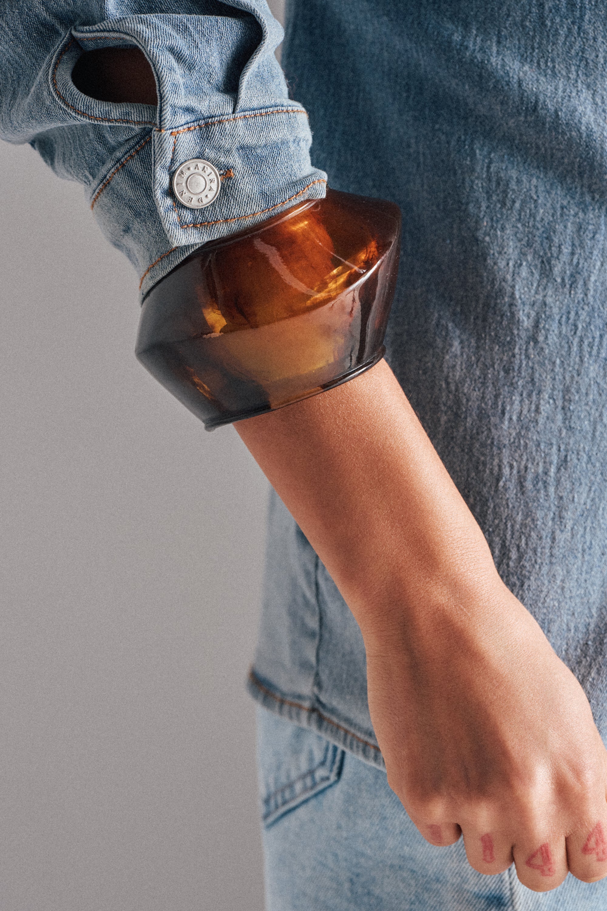 Wearing a rolled-up denim shirt, a person displays the IN THE SHOW BRACELET CUFF—a large, marbled amber accessory—on their wrist. The plain, neutral background highlights this statement piece.