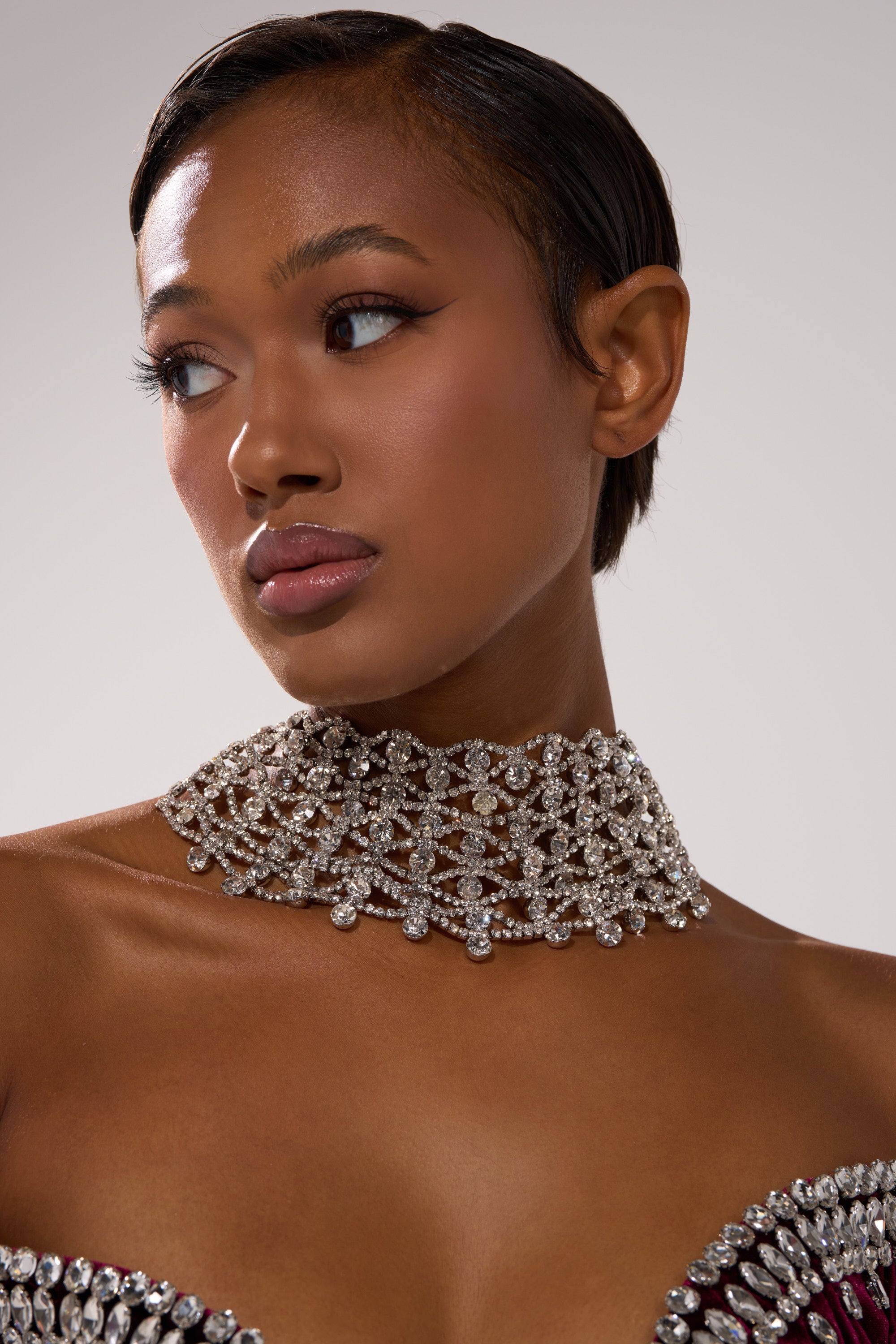 A woman with short hair looks to the side, wearing the FOR THE PARTY NECKLACE as statement jewelry. Her makeup is natural with winged eyeliner, and she pairs it with an elegant, strapless outfit.