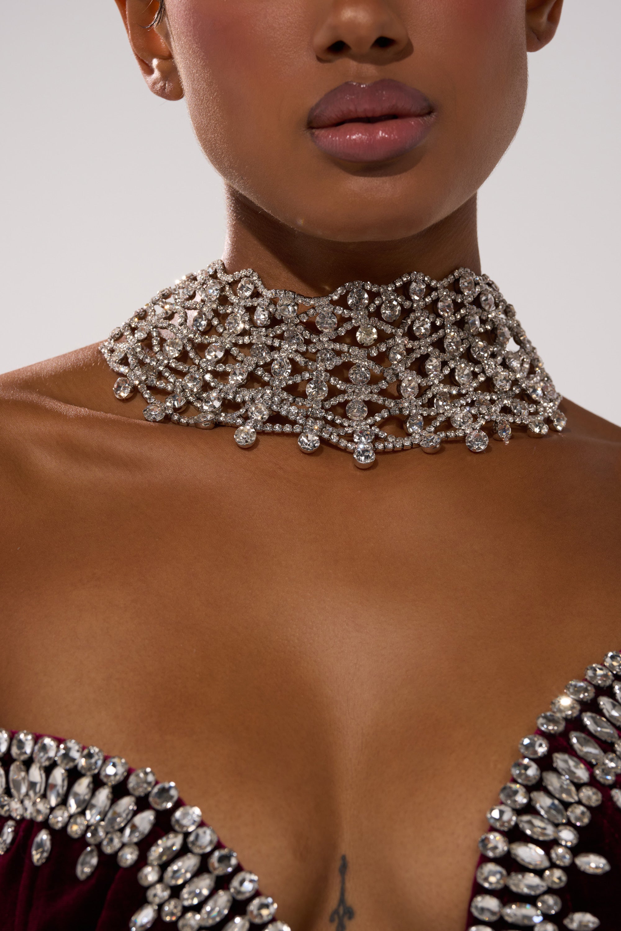 A close-up of someone wearing the FOR THE PARTY NECKLACE and a burgundy off-the-shoulder top with gemstone details. Their lips are slightly parted, and a small collarbone tattoo accentuates the bold statement jewelry.